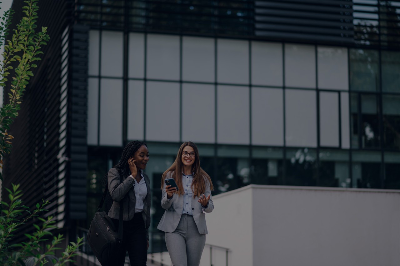 Young successful african american businesswoman in suit is talking on a smartphone while her caucasian female colleague is discussing success of their project. Buildings in the background. Copy space